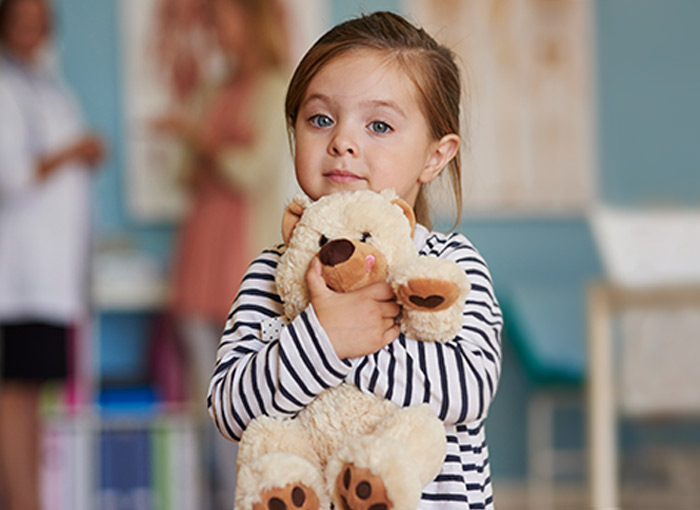 Young girl holds stuffed teddy bear, mom and doctor in white lab coat talk in background Young girl holds stuffed teddy bear, mom and doctor in white lab coat talk in background