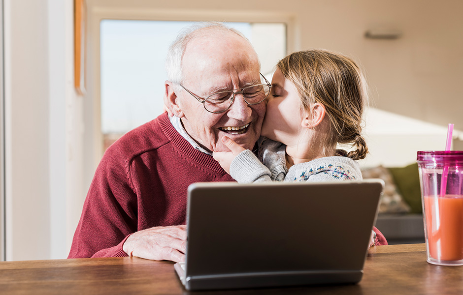 Little girl gives smiling grandfather a big kiss as he looks at laptop Little girl gives smiling grandfather a big kiss as he looks at laptop